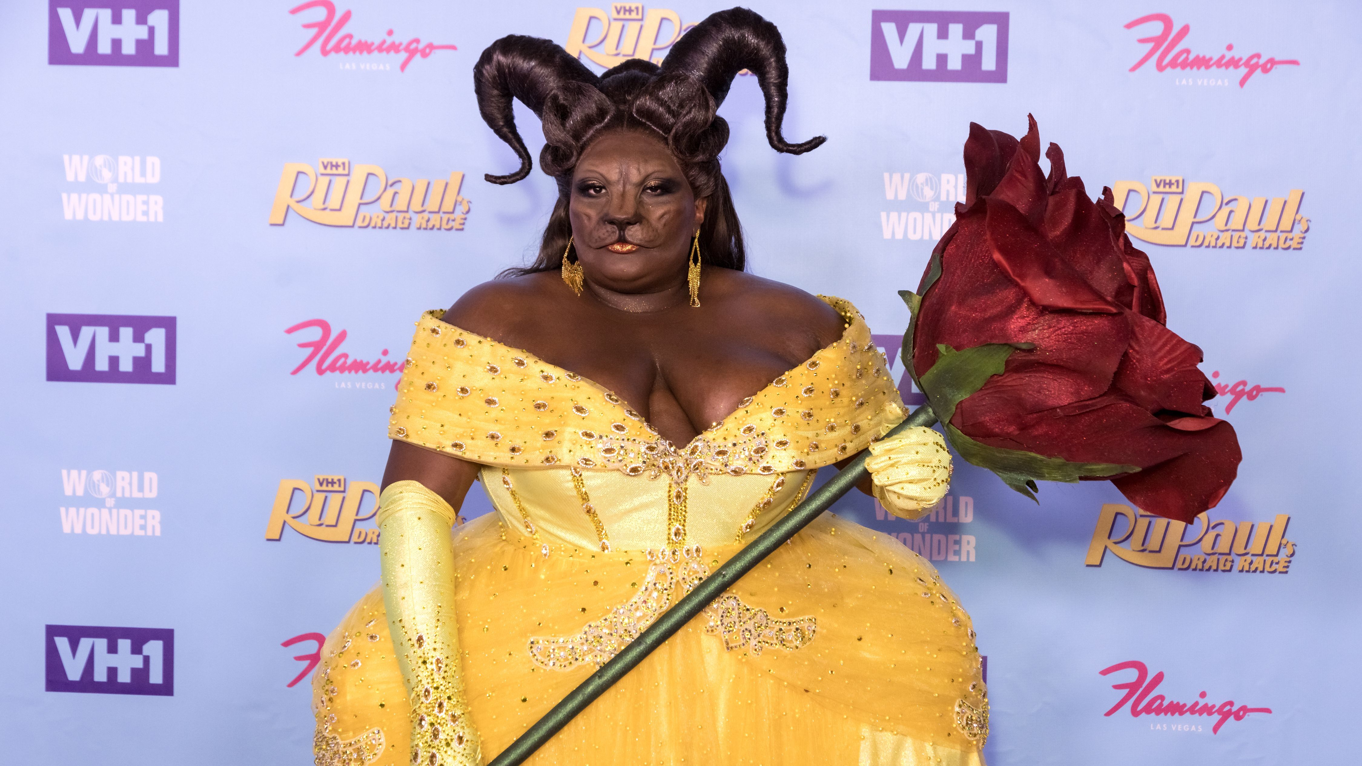 Cena da série RuPaul's Drag Race, mostra uma drag queen negra usando uma roupa inspirada na Bela e a Fera. Ela é uma mulher gorda, veste um vestido amarelo, segura uma rosa gigante e tem maquiagem com traços de besta, com chifres, nariz de fera e uma peruca que imita chifres. 