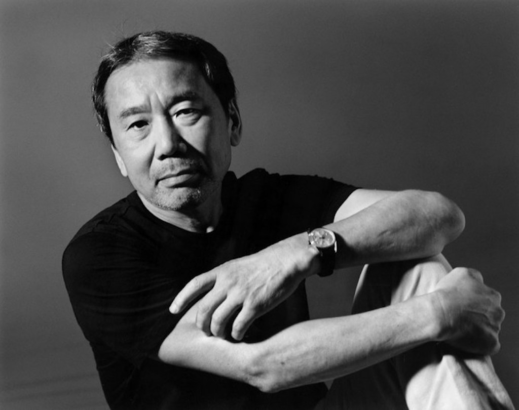 Foto em preto e branco do escritor japonês Haruki Murakami. Ele é um homem com traços asiáticos, possui cabelo e barba grisalhos, e na foto está sentado, olhando diretamente para a câmera, com a mão esquerda sobre o braço direito e seu braço direito apoiado sobre a perna esquerda. Ele veste uma camiseta preta e uma calça cinza. O fundo da imagem também é cinza.