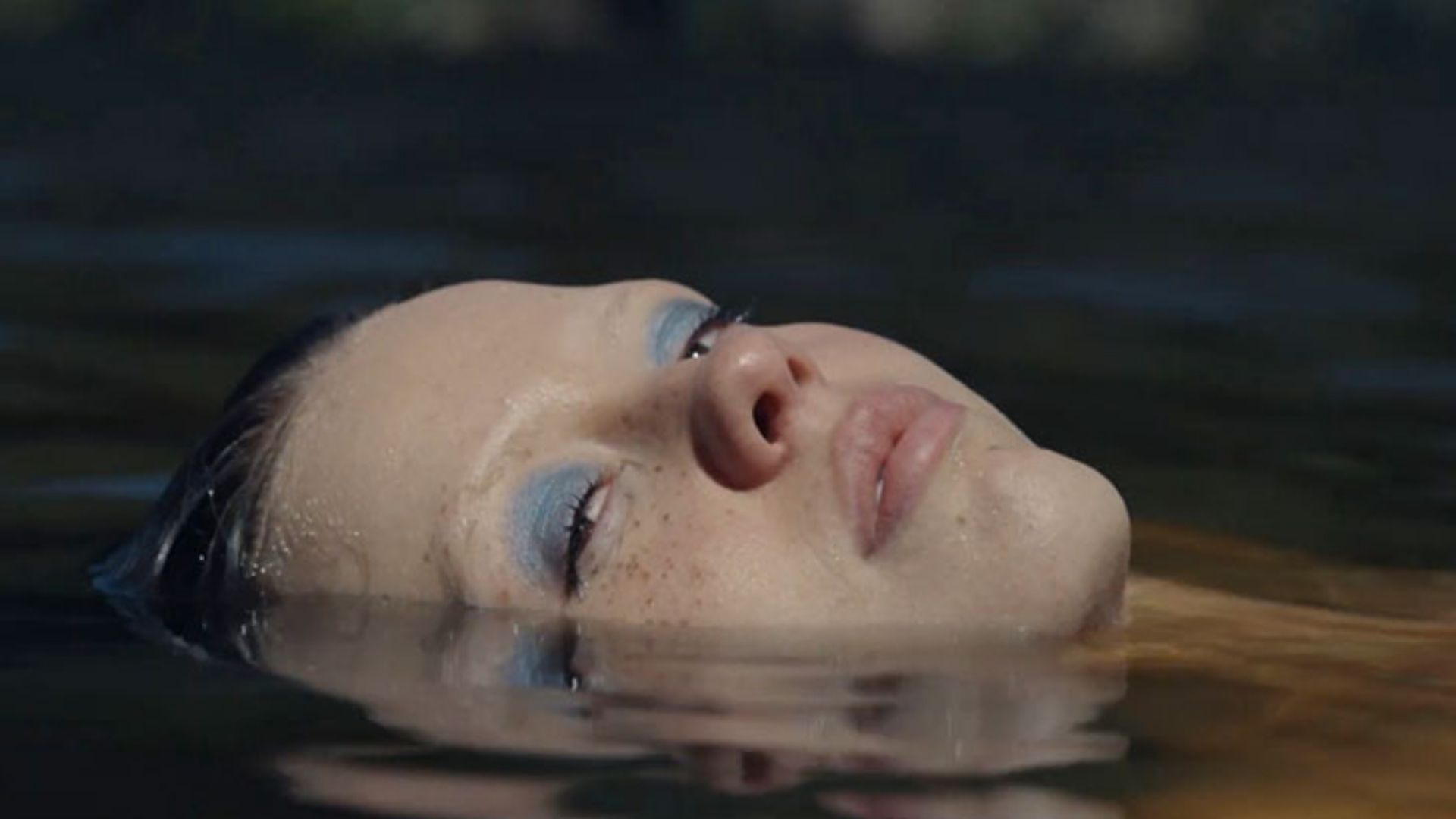 Cena do filme X, mostra uma mulher branca flutuando deitada num rio, com apenas a cabeça para fora da água.