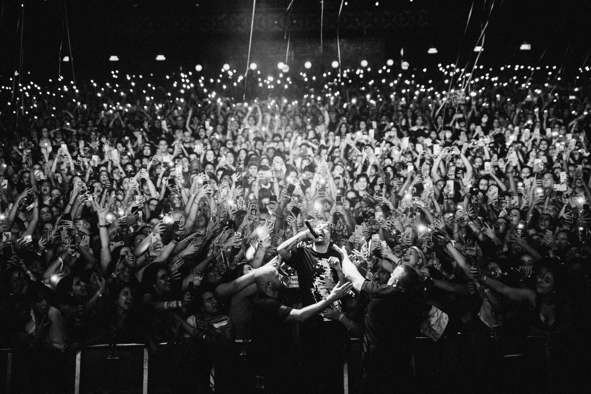A imagem mostra o cantor Louis Tomlinson, homem branco de cabelos curtos, que usa uma blusa preta e tem um microfone na mão esquerda, se jogando em meio a uma plateia de milhares de pessoas. A foto está em preto e branco e conseguimos ver várias luzes refletidas ao fundo.