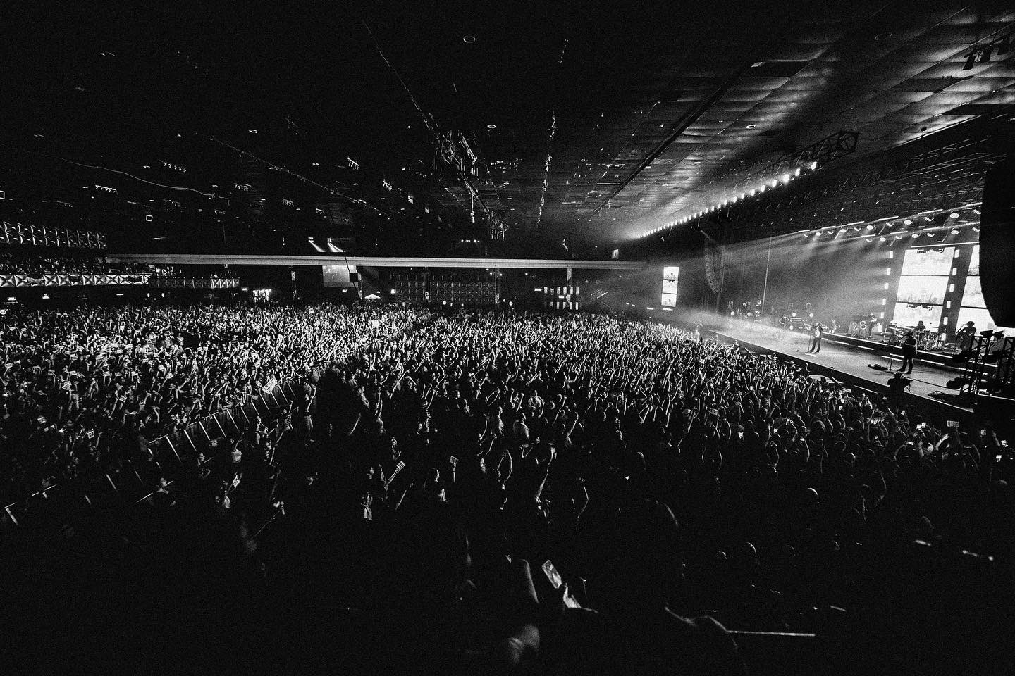 A imagem mostra uma multidão de pessoas durante o show de Louis Tomlinson. No canto direito vemos o palco, onde o cantor e sua banda estão posicionados. A imagem é preta e branca.