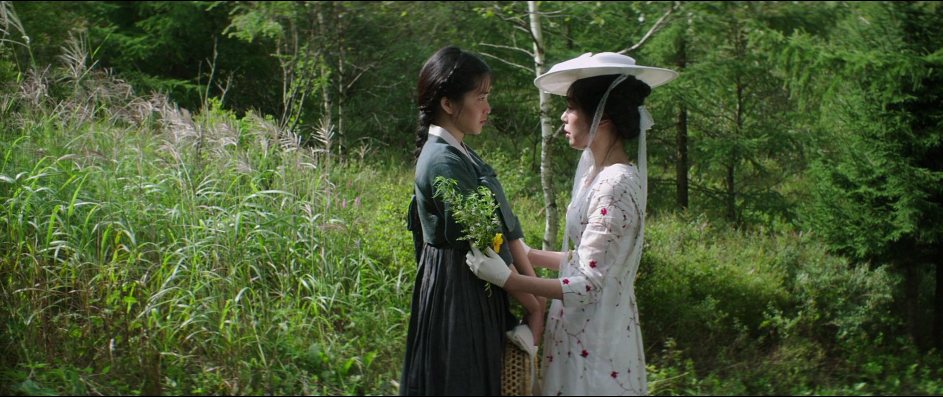 Cena do filme A Criada. Na imagem da esquerda para a direita, Sook-hee e Hideko, duas mulheres asiáticas de cabelos escuros. Sook-hee veste cinza e Hideko veste branco. Hideko segura Sook-hee pelos braços. Ao fundo, a mata verde com flores.