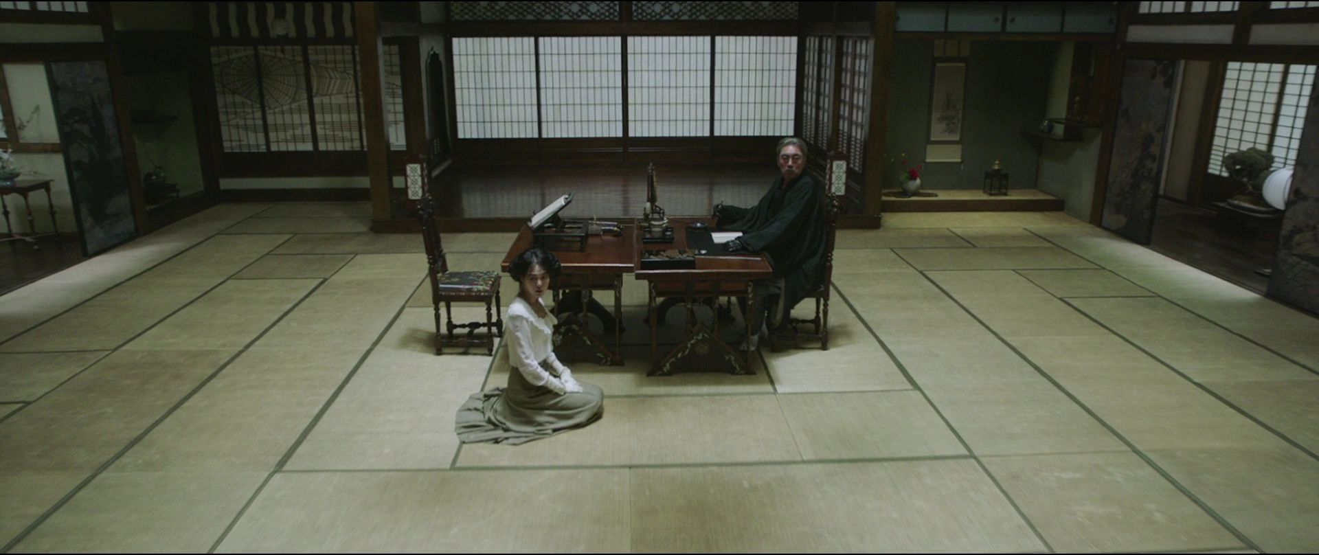 Cena do filme A Criada. Na imagem da esquerda para a direita, Hideko, uma mulher asiática de cabelos escuros está sentada ao pé do seu tio Kouzuki. Ele é um homem asiático de cabelos grisalhos. Ela veste branco e ele veste preto. O cenário é uma biblioteca.