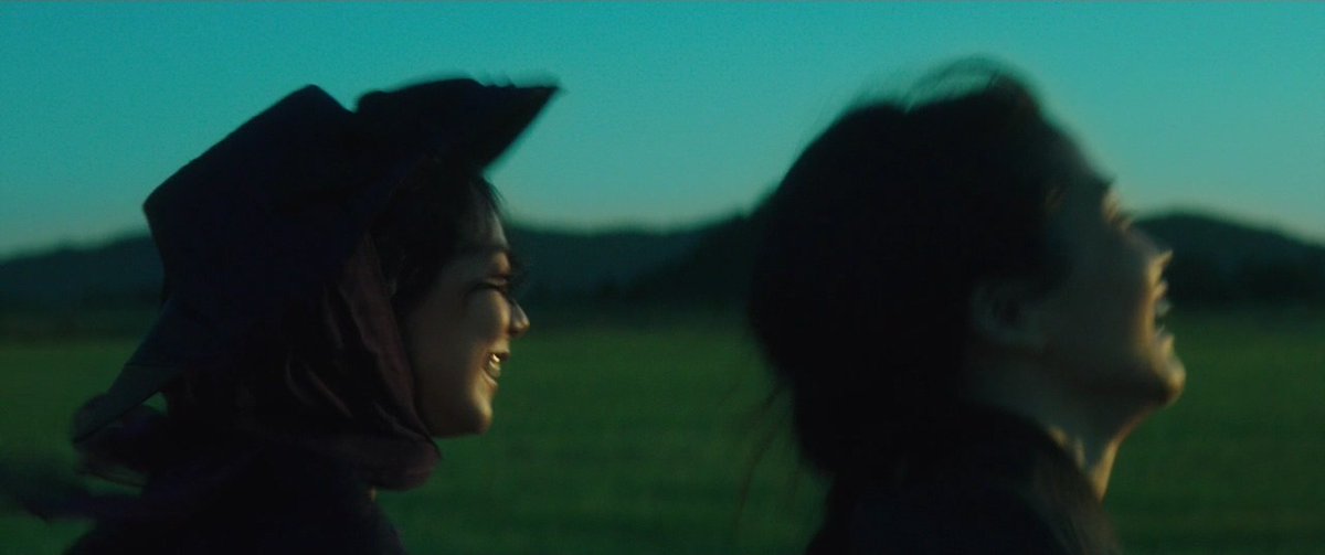 Cena do filme A Criada. Na imagem da esquerda para a direita, Hideko e Sook-hee, duas mulheres asiáticas de cabelos escuros, correm enquanto sorriem. Ambas vestem preto. Ao fundo, uma imensidão verde.