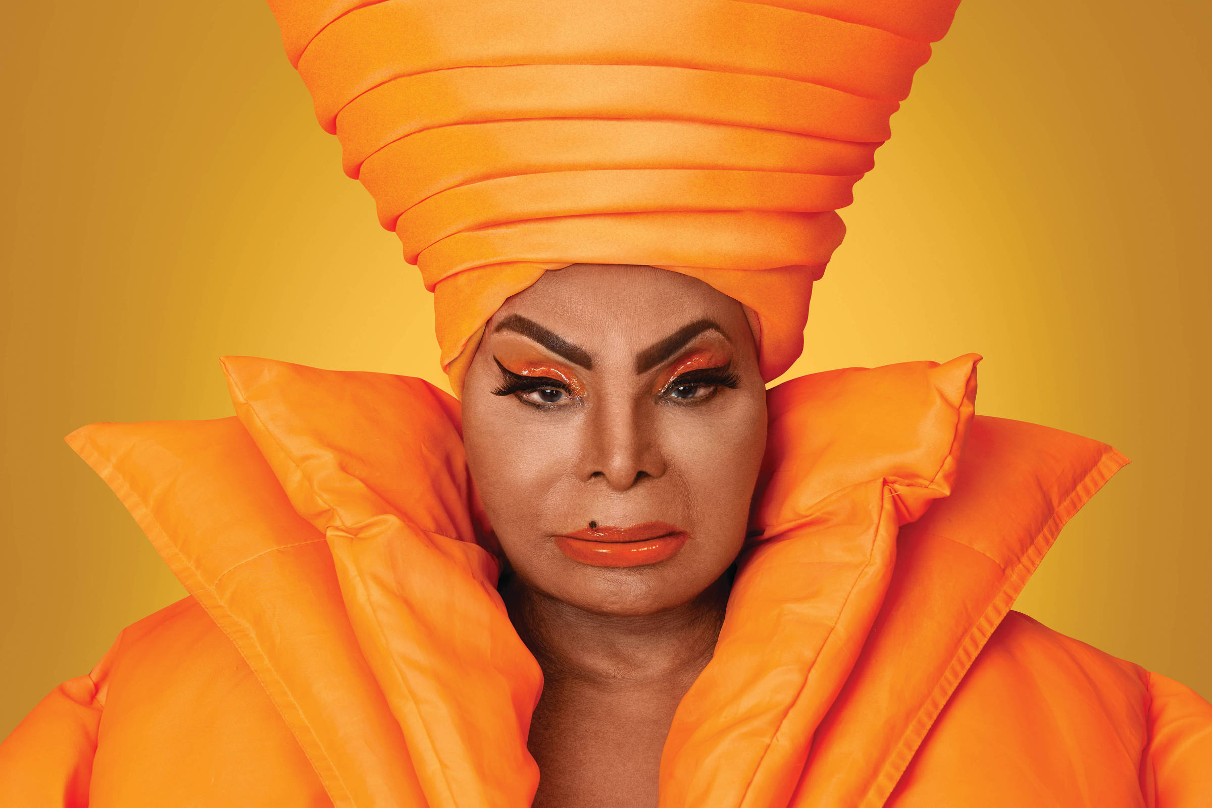 Retrato da cantora Elza Soares. Fotografia retangular horizontal, com fundo alaranjado. A artista brasileira ocupa quase toda a imagem. Ela é uma mulher negra, maquiada, olha para frente e veste roupas laranjas, com destaque para um grande turbante na cabeça.