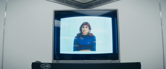 Cena de Severance. Nela, Helly, uma mulher branca de cabelos ruivos aparece em uma tv antiga. Ela veste um vestido azul e a tv está sobre um carrinho em uma sala branca.