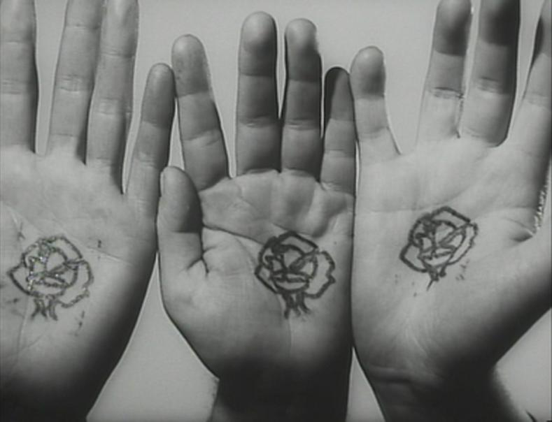  Cena do filme O Funeral das Rosas. A imagem em preto e branco mostra três mãos humanas com as palmas voltadas para frente. Em cada palma há o desenho de uma rosa.