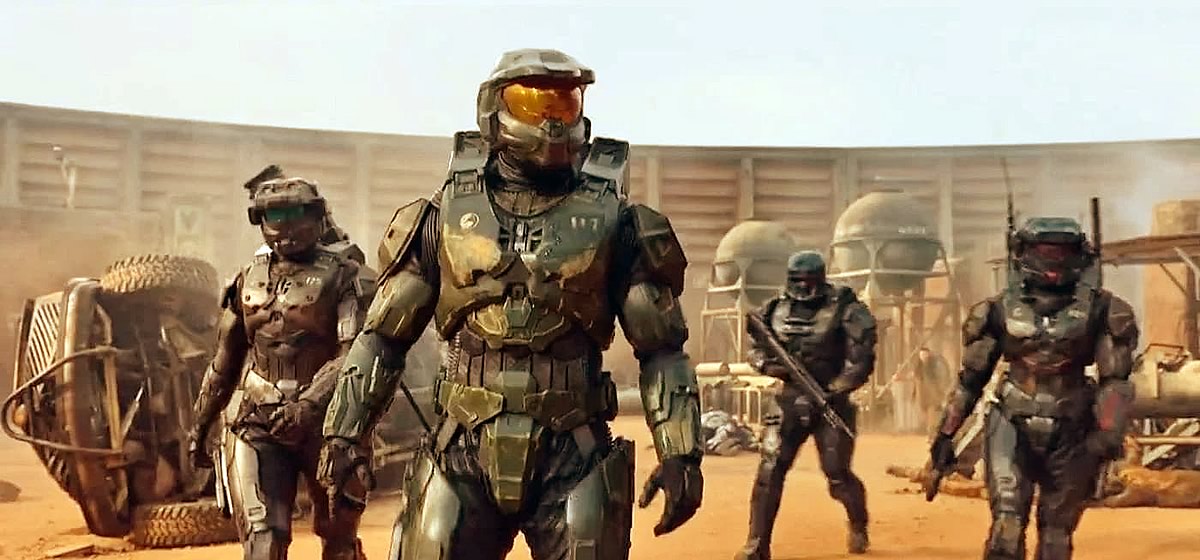 Cena da série Halo exibe um grupo de quatro soldados em um ambiente árido, cercado por um muro alto. Todos vestem uma armadura com capacete. A armadura do líder do grupo, à frente, é verde e o visor do capacete é dourado. A armadura dos soldados ao redor é preta e todos estão armados. 