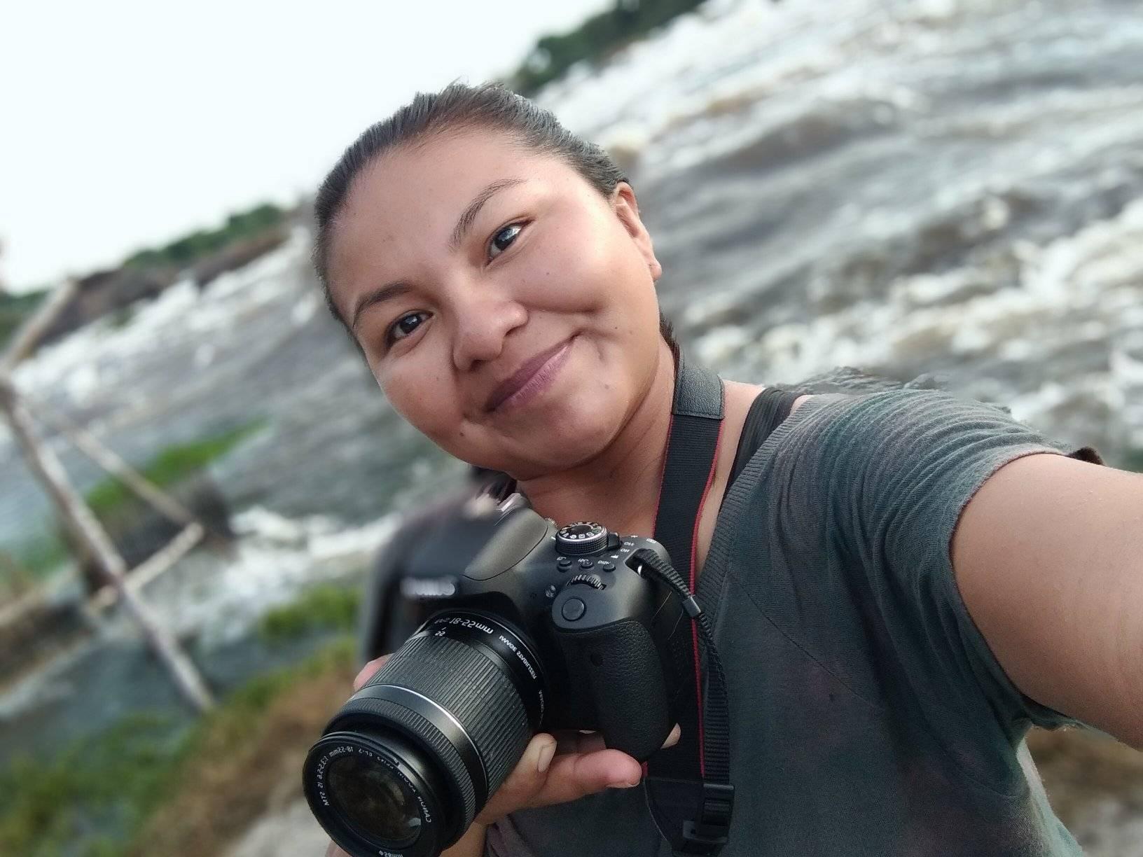  Selfie tirada pela cineasta Larissa Ye`padiho Duarte Tukano, mulher indígena do povo Tukano. Ela olha diretamente para câmera e sorri sem mostrar os dentes enquanto segura uma câmera fotográfica. Ela veste uma camiseta na cor cinza e está numa paisagem com o Rio Negro desfocado.