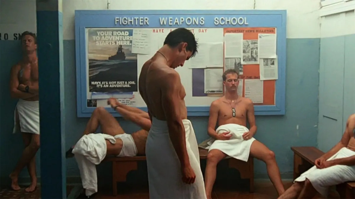 Cena do filme Top Gun, mostra um vestiário masculino cheio de homens brancos com toalhas amarradas na cintura. 
