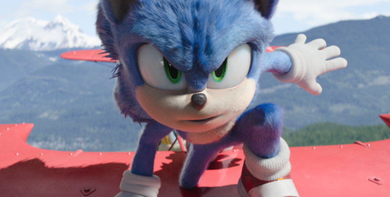 Cena do filme Sonic 2. Sonic, uma criatura azul e de olhos verdes, que se parece com um ouriço, se posiciona em cima das asas de uma nave vermelha. Ao fundo existem montanhas com neve e mata.