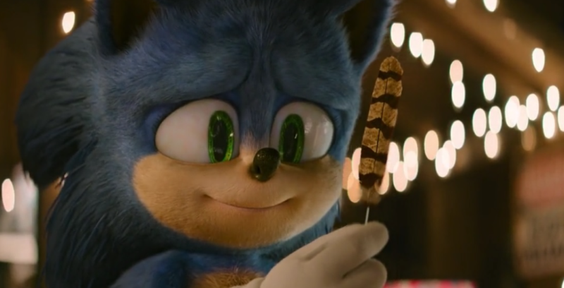 Cena do filme Sonic 2. Sonic olha para uma pena de ave, com listras marrons e bege. Ele tem uma expressão de carinho no rosto e um pequeno sorriso. Ao fundo está seu quarto, embaçado pelas luzes do Sol