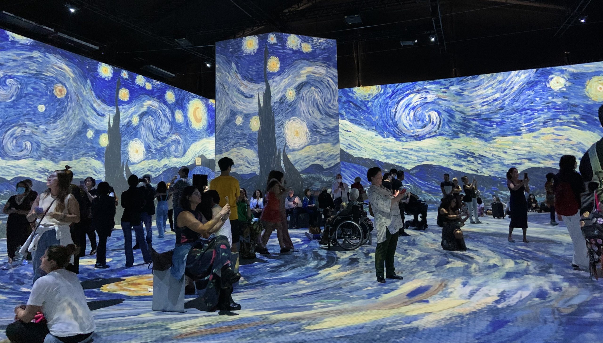 Imagem da sala principal projetando uma das obras mais famosas de Van Gogh, A Noite Estrelada, e várias pessoas prestigiando a apresentação, tanto em pé quanto sentadas, no chão ou em bancos.