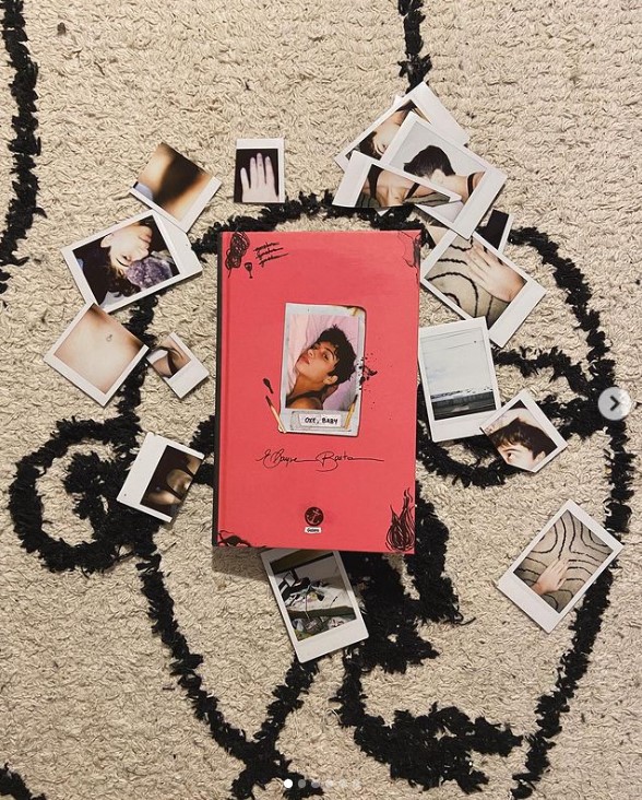 Fotografia quadrada do segundo livro publicado por Elayne. A capa do livro é rosa, tem uma Polaroid no meio com uma foto da autora, ao lado tem alguns fósforos e detalhes em preto. O fundo é de um tapete preto e branco, além de outras Polaroids no chão perto do livro. 