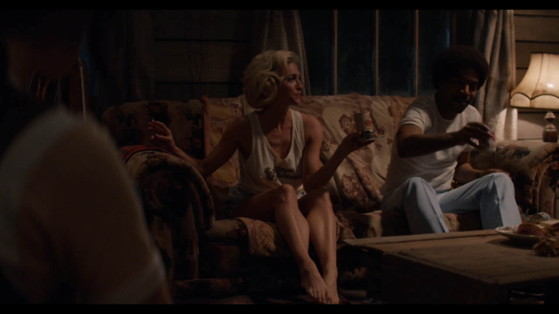 Cena do filme X. Na cena, em uma cabana pouco iluminada, vemos uma mulher loira, aparentando cerca de 35 anos, sentada em um sofá. Ao lado direito dela, vemos um homem negro, aparentando cerca de 35 anos.