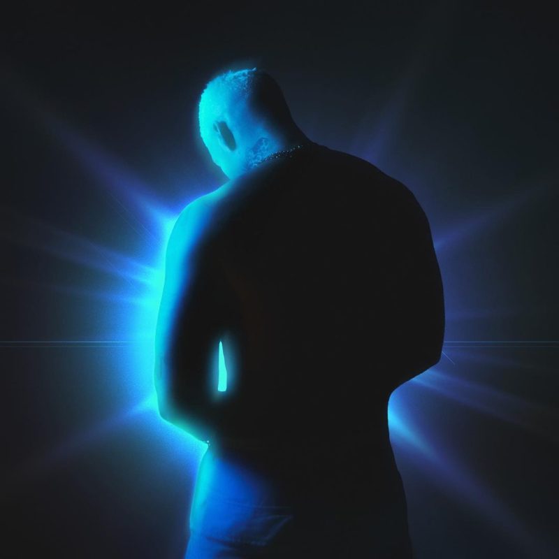 A foto mostra Filipe Ret de costas, em um fundo preto azulado. Há uma luz azul que contorna o corpo de Ret.