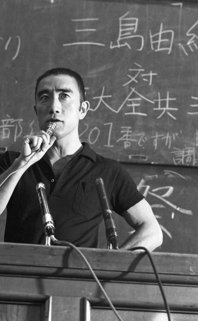 Yukio Mishima segurando um microfone enquanto discursa em um palanque, em frente a um quadro negro, com uma expressão austera