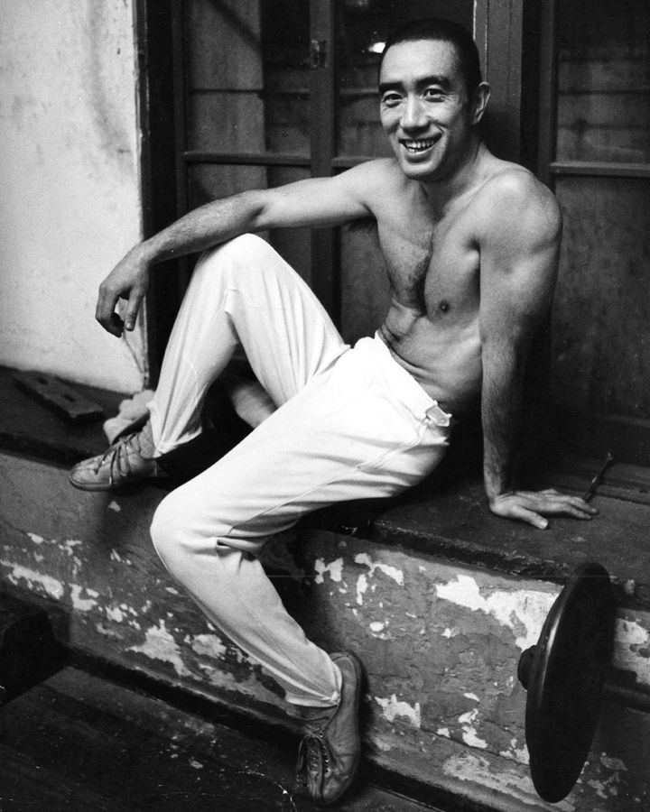O autor Yukio Mishima sorri sem camisa sentado em um balcão, enquanto veste uma calça branca e revela o seu porte físico