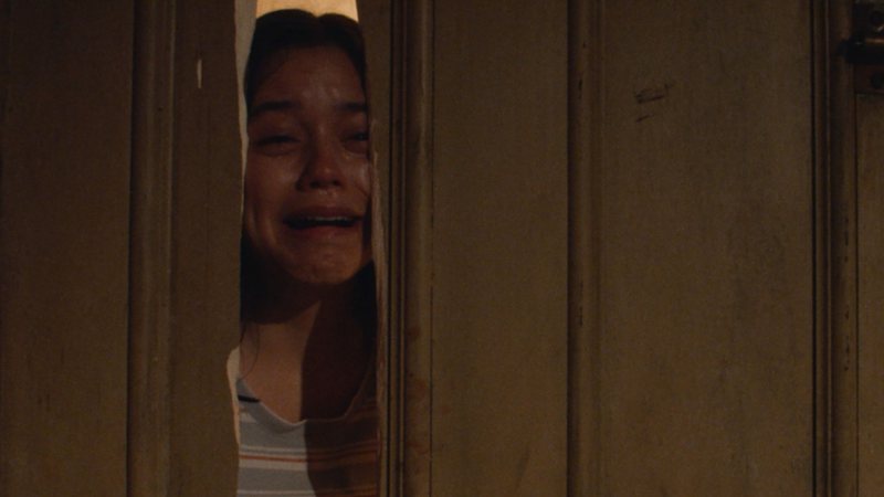 Cena do filme X. Na cena, por uma fresta de uma porta quebrada, vemos uma mulher, aparentando cerca de 18 anos, chorando.