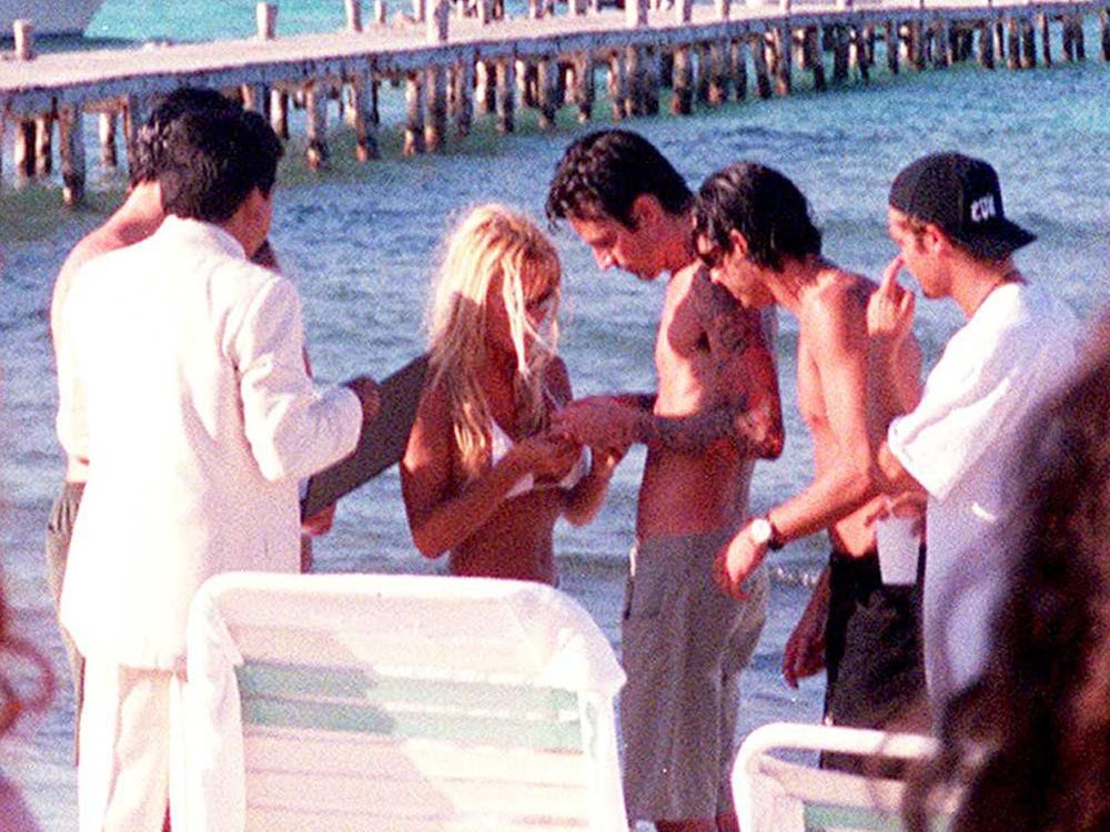 Foto do casamento de Pamela Anderson e Tommy Lee. Na fotografia, vemos pessoas em um primeiro e segundo plano, e a praia ao fundo. Ao centro da imagem, vemos Pamela Anderson, à esquerda, e Tommy Lee, à direita. Ela veste um biquíni branco e ele veste somente uma bermuda cinza. Ambos olham para algo em suas mãos.