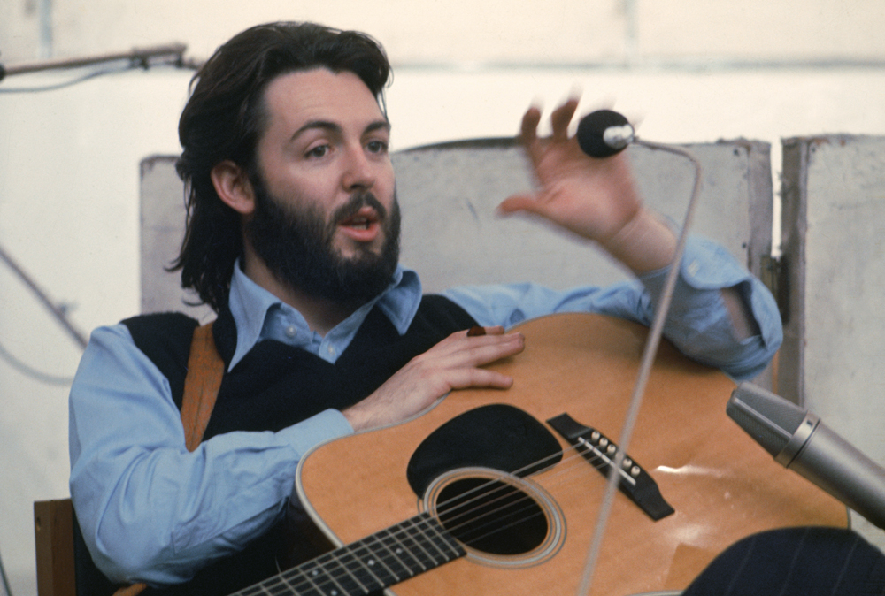 Cena do documentário The Beatles: Get Back exibe Paul McCartney, um músico branco, com cabelo comprido e barba cheia, sentado com seu violão no colo. Ele veste uma camisa azul com um suéter preto por cima e gesticula, como se estivesse explicando algo a alguém fora de cena. 