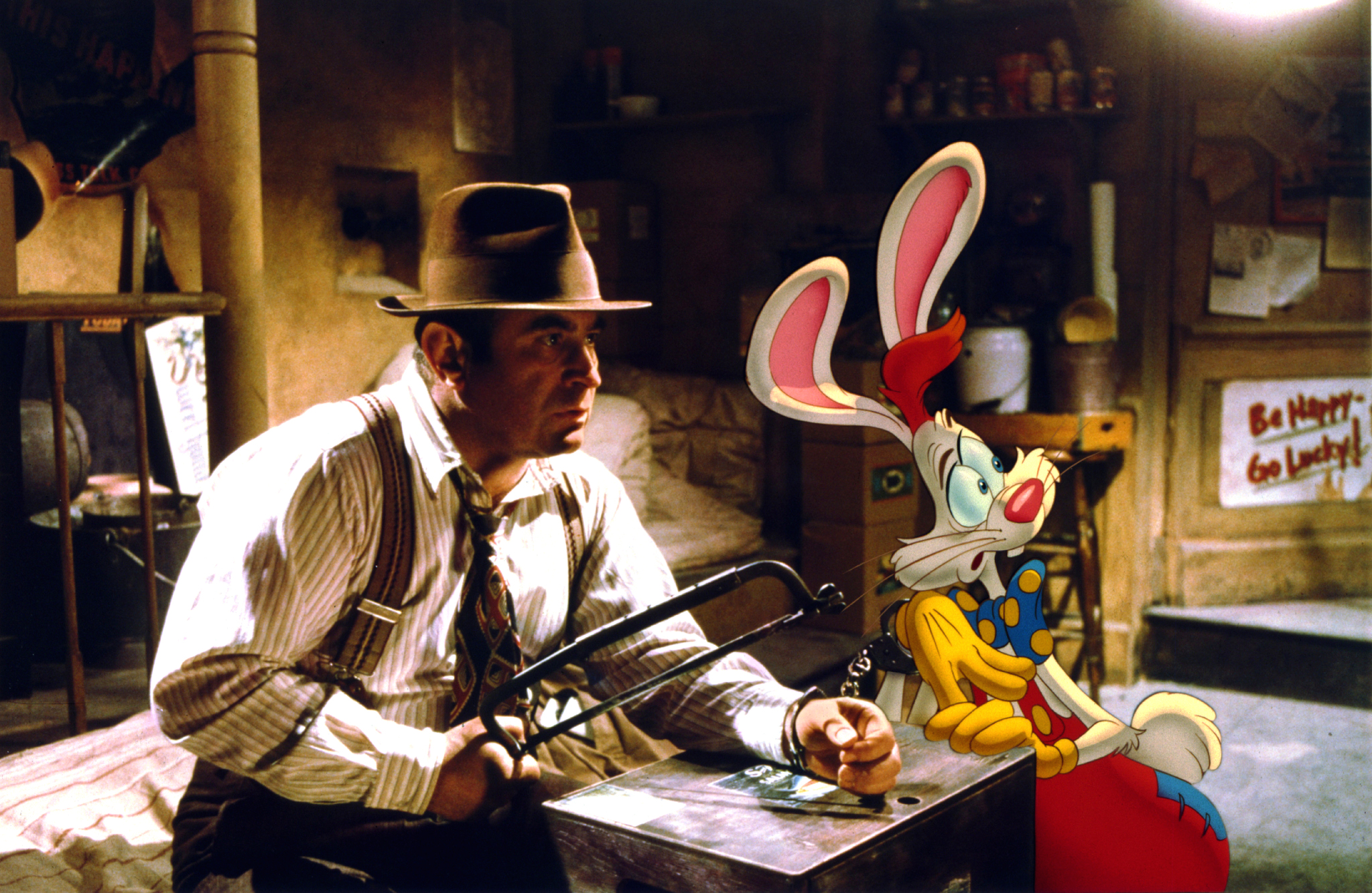 Cena do filme Uma Cilada para Roger Rabbit. No lado esquerdo da imagem, temos o ator Bob Hoskins, um homem branco e baixinho de chapéu marrom vestindo camisa social branca listrada, suspensório marrom, gravata com detalhes em azul e vermelho e uma serradura na mão esquerda, com a mão direita estendida sobre uma pequena mesa marrom escura. A mão direita está algemada a um coelho que se encontra no lado direito da imagem. O coelho Roger Rabbit é um coelho branco de grandes orelhas rosadas, olhos azuis, focinho também rosado, gravata borboleta azul com bolinhas amarelas, luvas amarelas e macacão vermelho. A cena acontece dentro de um quarto abandonado durante a noite.