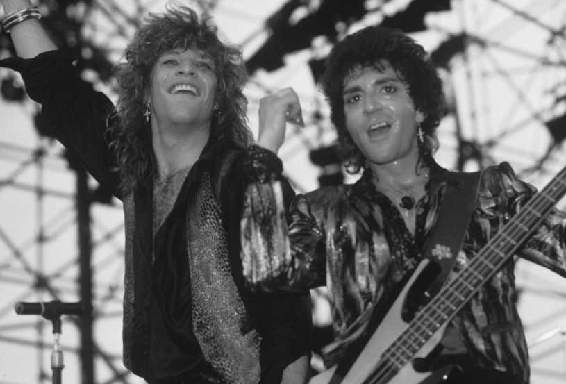  Fotografia de Jon Bon Jovi e Alec John Such nos palcos. Jovi e Such são dois homens brancos de cabelos compridos. Na esquerda, Jon Bon Jovi aparece sorrindo para a plateia, enquanto na direita, Alec John Such toca baixo e também sorri.