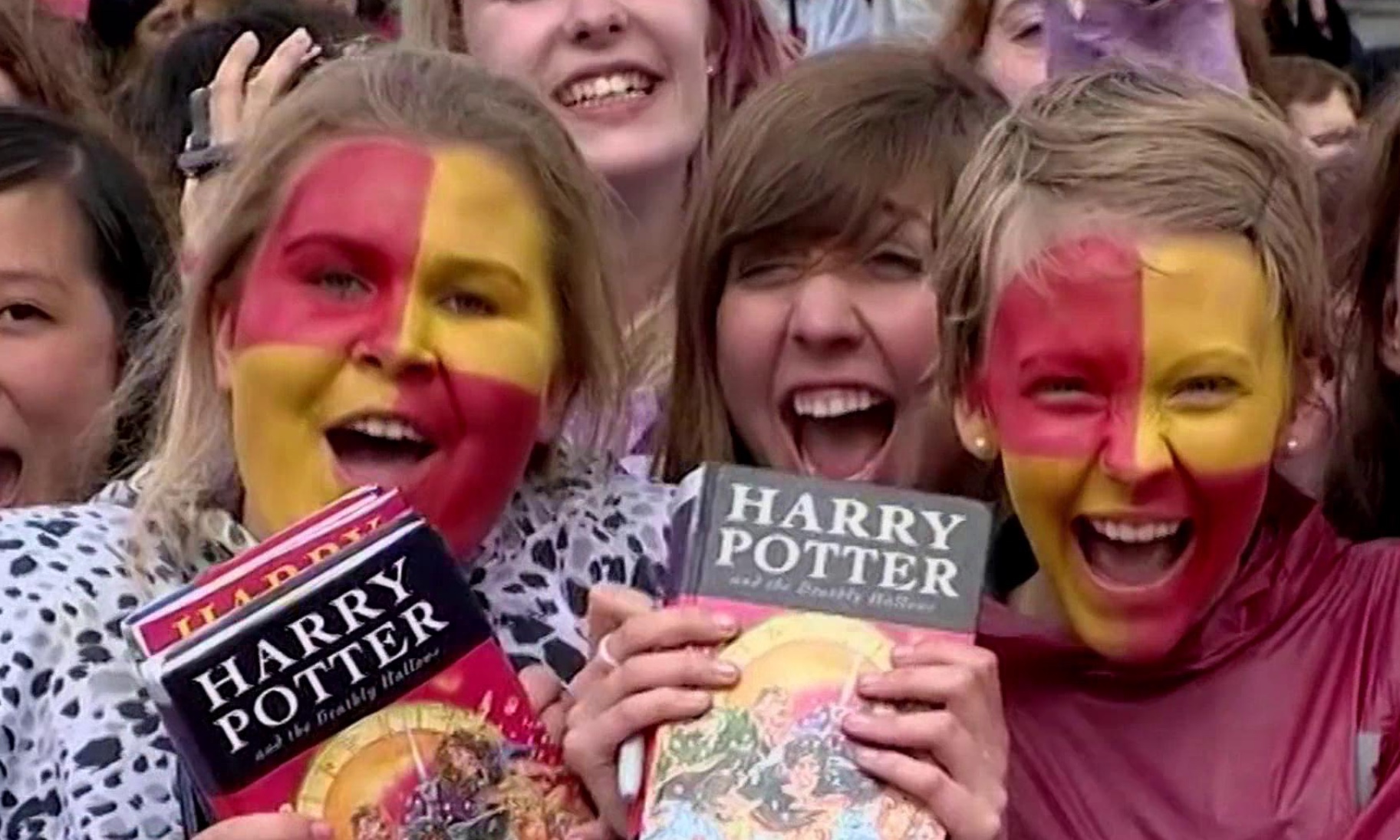 Cena do documentário Harry Potter 20th Anniversary: Return To Hogwarts exibe um grupo de fãs de Harry Potter. Em destaque estão duas meninas brancas e loiras com o rosto pintado de vermelho e amarelo, com expressão de animação segurando um exemplar do livro Harry Potter e as Relíquias da Morte.