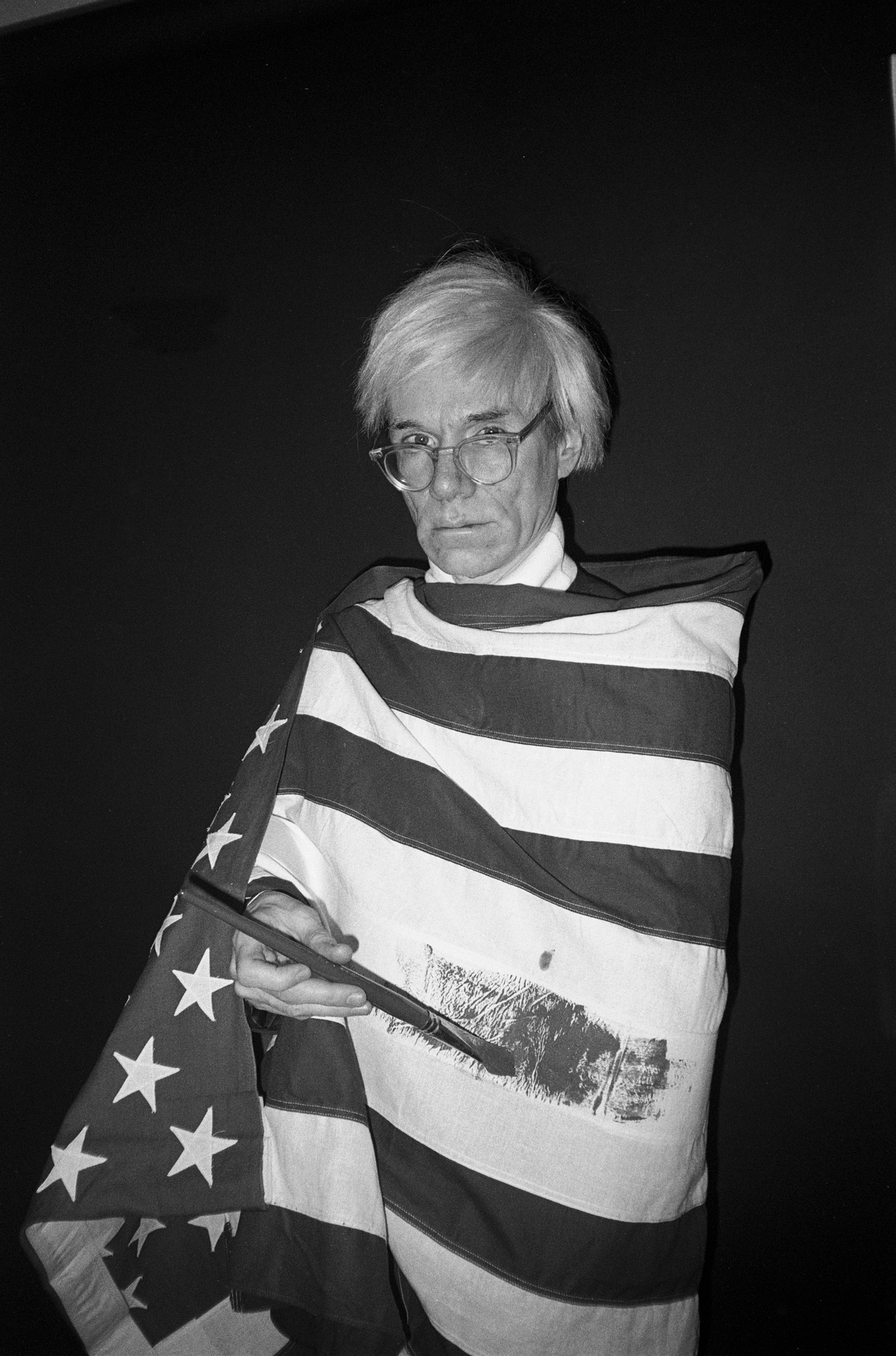foto preto e branco de Andy Warhol, enrolado na bandeira dos Estados Unidos. Usa óculos redondos de arame e sua característica peruca loira, enquanto sua mão, segurando um grande pincel preto, pinta outra faixa, provavelmente azul, na parte branca da bandeira. O fundo é escuro.