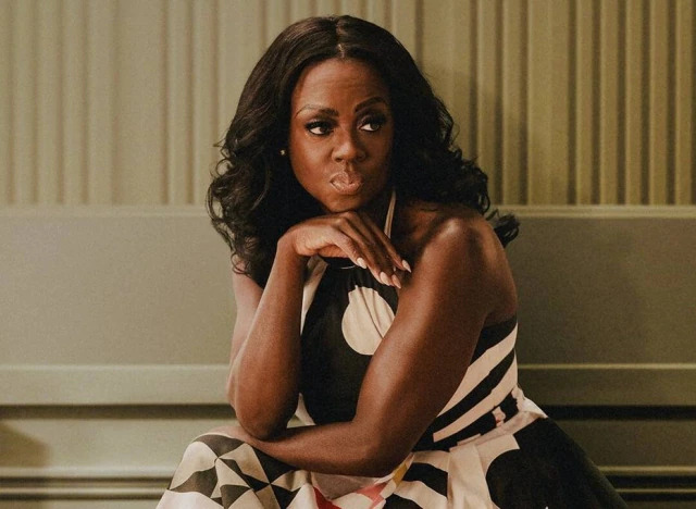 Foto da série The First Lady. Na imagem está a personagem Michelle Obama, interpretada pela atriz Viola Davis. Michelle é uma mulher negra, tem cabelos pretos ondulados de comprimento médio e está usando um vestido frente única com estampas geométricas em preto e branco. Ela está sentada com uma mão sob o queixo e a outra segurando a perna, seu rosto é sério com um leve bico nos lábios. Ao fundo, há uma parede branca e bege com textura listrada.