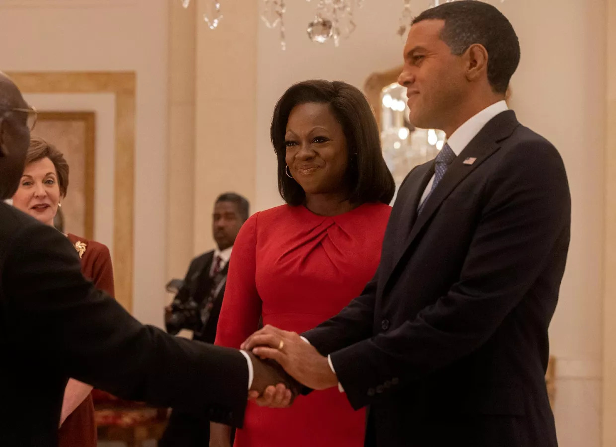 Foto da série The First Lady. Na imagem estão os personagens Michelle Obama, interpretada por Viola Davis, e Barack Obama, interpretado por O-T Fagbenle. Michelle é uma mulher negra de cabelos pretos curtos, está vestindo um vestido vermelho de manga 3/4 e está sorrindo com os lábios fechados. Barack é um homem negro com cabelos escuros raspados, ele veste um terno preto com uma gravata azul. Os dois estão lado a lado, enquanto ele cumprimenta alguém com um aperto de mão. No plano de fundo, aparece um homem negro e uma mulher branca próximos a uma parede branca com detalhes dourados