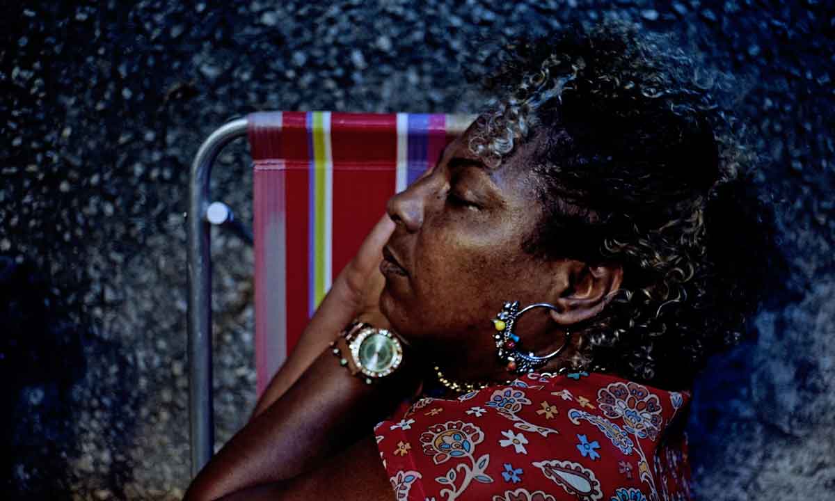 Cena do filme Marte Um, a imagem mostra uma mulher adulta negra dormindo de lado numa cadeira de praia vermelha. Ela usa brincos e relógio brilhantes e uma camiseta vermelha com detalhes em branco e azul.