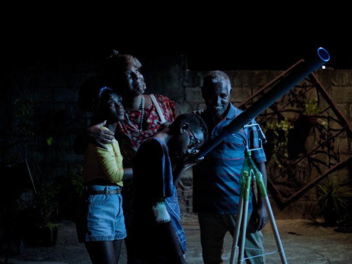 Cena do filme Marte Um, a imagem mostra quatro pessoas negras da mesma família de pé e reunidas ao redor de um telescópio. Está de noite e a luz azul da lua reflete neles.