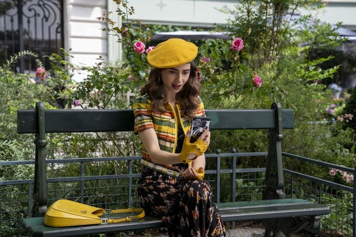 Emily está sentada num banco vestindo um vestido preto florido e uma camisa de mangas curtas com listras amarelas. Ela usa acessórios amarelos também, como boina e bolsa. Ao fundo vê-se alguns arbustos com flores cor-de-rosa.