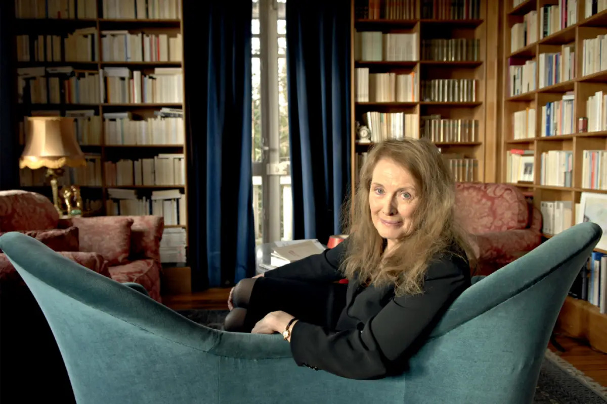 Fotografia de Annie Ernaux. A autora está ao centro, num sofá azul. Ela usa uma camisa preta, seus cabelos são claros e lisos. Annie sorri levemente para a foto. Ao fundo, existe uma estante de livros. 