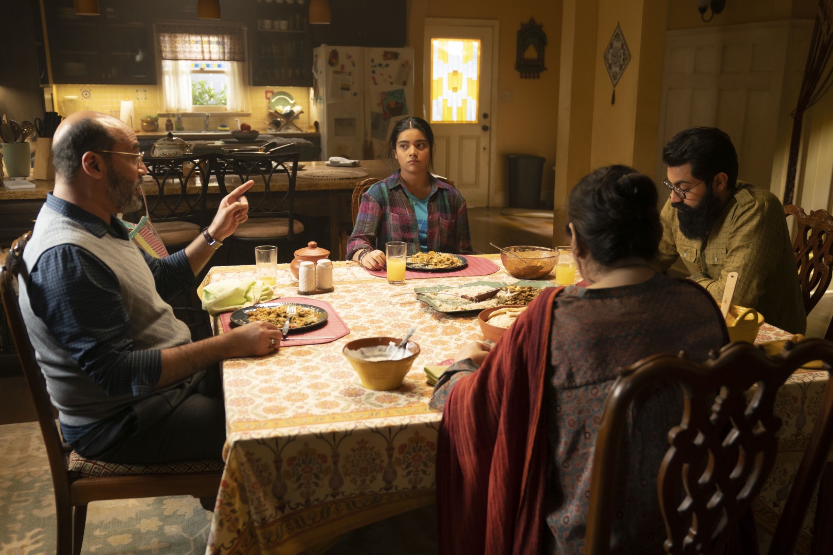 Cena da primeira temporada de Ms. Marvel. A família Khan come uma refeição sentados na mesa de jantar de sua residência. Vemos Kamala (Iman Vellani) na lateral superior da mesa quadrada, olhando emburrecida para seu pai, Yusuf (Mohan Kapur), à esquerda. Vemos apenas as costas de sua mãe, Muneeba (Zenobia Shroff), que se senta na lateral inferior da mesa, e apenas parte do perfil de seu irmão mais velho, Aamir (Saagar Shaikh), na lateral direita, olhando para seu prato. Kamala é uma menina paquistanesa de pele marrom, com cabelos pretos e longos, amarrados para trás num rabo de cavalo, usando uma camisa xadrez vermelha por cima de uma camiseta azul bebê. Yusuf é um homem paquistanês de pele marrom, cabelos e barba grisalhos e começando a ficar calvo, usando uma camisa xadrez azul escura por cima de um suéter cinza sem mangas. Muneeba é uma mulher paquistanesa de pele marrom, com os cabelos pretos presos num coque, usando um vestido tradicional rubro e azul com um tecido vermelho apoiado no seu ombro esquerdo. Aamir é um homem paquistanês de pele marrom, com cabelos pretos e uma barba longa e espessa, usando um par de óculos e uma camisa xadrez verde clara abotoada. A mesa está coberta por um tecido amarelo e branco e, acima dele, os Khan comem em seus pratos, com diversos condimentos espalhados pelos cantos da mesa e um prato principal no centro. Atrás deles, podemos ver a cozinha da casa, com uma geladeira coberta de imãs no canto direito, ao lado de uma porta branca com vitrais amarelos e azuis. As paredes da casa são amarelas e o chão é de madeira, com um tapete cinzento cobrindo a sala de jantar onde a mesa se encontra.