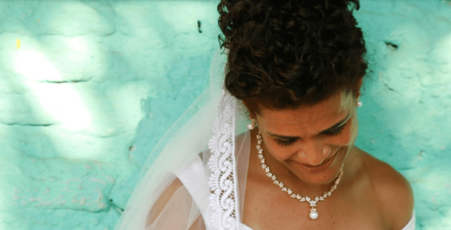 Cena do filme Paloma, mostra uma mulher negra sorrindo e olhando para baixo. Ela está à frente de uma parede azul e usa roupas de casamento. Um vestido branco de noiva, com véu e colar na mesma cor.