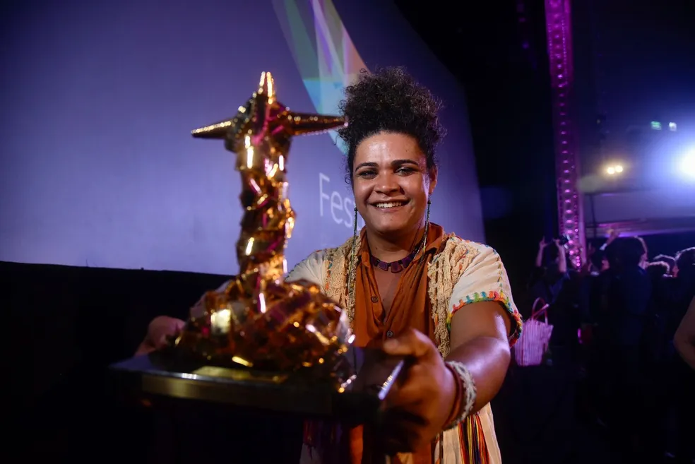 Foto da atriz Kika Sena, uma mulher negra, segurando o troféu de Melhor Atriz no Festival do Rio. Ela usa roupas claras com um lenço marrom em volta do pescoço e sorri. O troféu é cor de cobre e tem o formato parecido com o do Cristo Redentor.