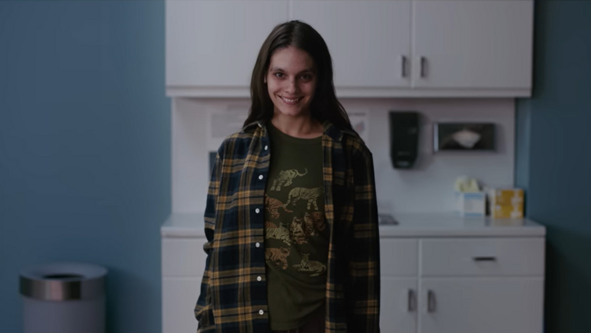 Cena do filme Sorria, mostra uma jovem branca parada e olhando para a câmera, sorrindo de maneira assustadora. Ela usa blusa verde e camisa xadrez em tons terrosos. 