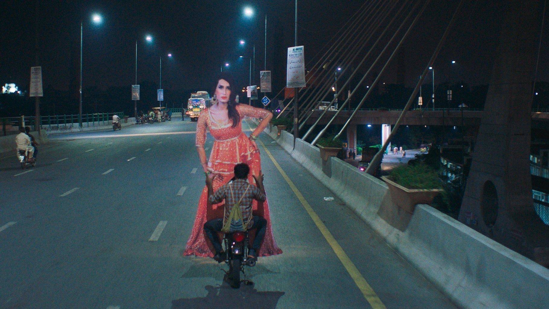 Cena do filme Joyland, mostra uma estrada de noite com um homem andando de moto, de costas para a câmera. Ele leva um papelão recortado no formato de uma mulher, usando roupas vermelhas e com a mão na cintura.
