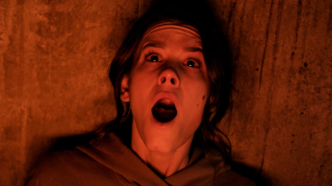 Cena do filme Sorria, mostra uma mulher branca encostada na parede, com expressão de medo no rosto, a boca aberta e os olhos lacrimejando, sendo iluminada por uma luz laranja.