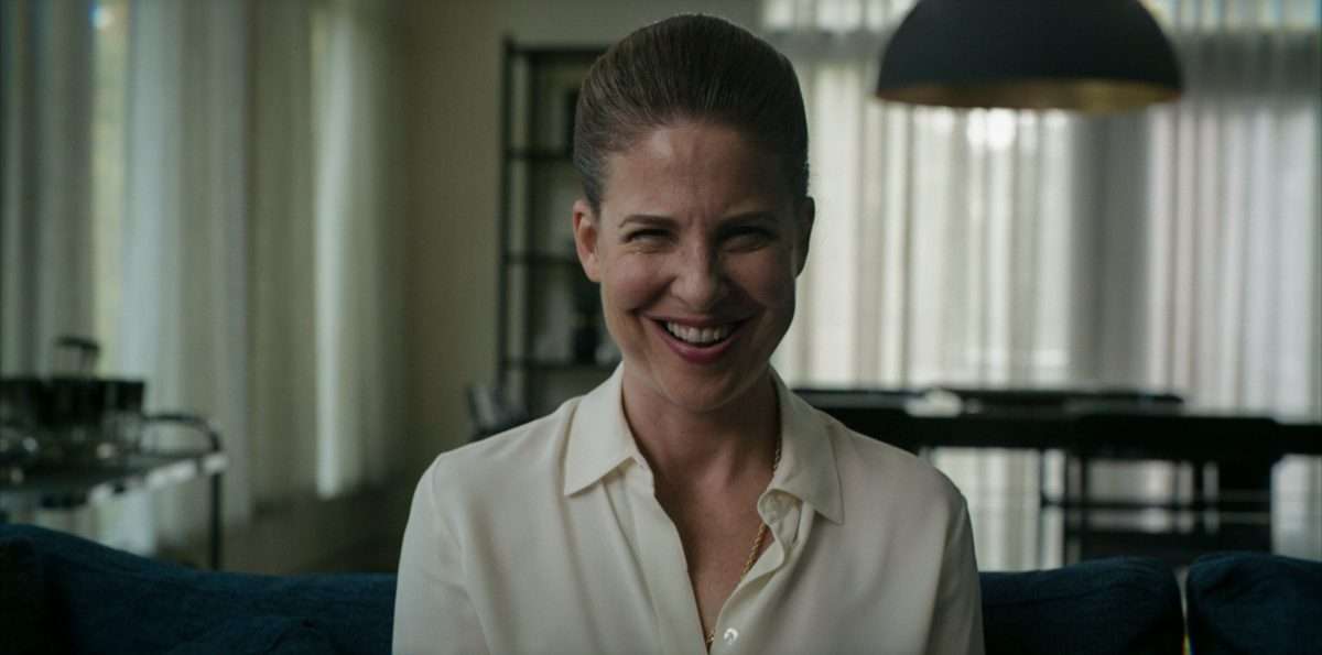 Cena do filme Sorria, mostra uma mulher branca sorrindo de maneira assustadora. Ela usa roupas brancas e olha em direção à câmera.