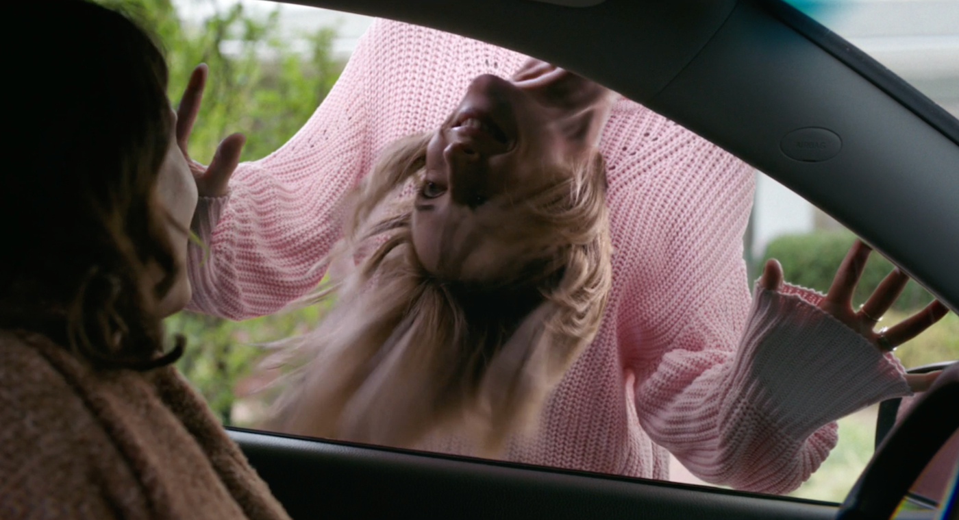 Cena do filme Sorria, mostra uma mulher dentro do carro, olhando de lado pela janela do motorista. Lá fora, vemos outra mulher, vestindo uma blusa rosa e com a cabeça caindo do pescoço, como um pêndulo, sorrindo de maneira assustadora.