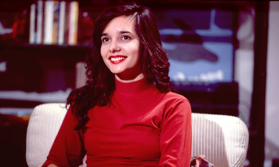 Temos na imagem Daniella Perez em uma entrevista para a rede Globo. A atriz usava uma blusa vermelha e esta sentada em uma poltrona branca.