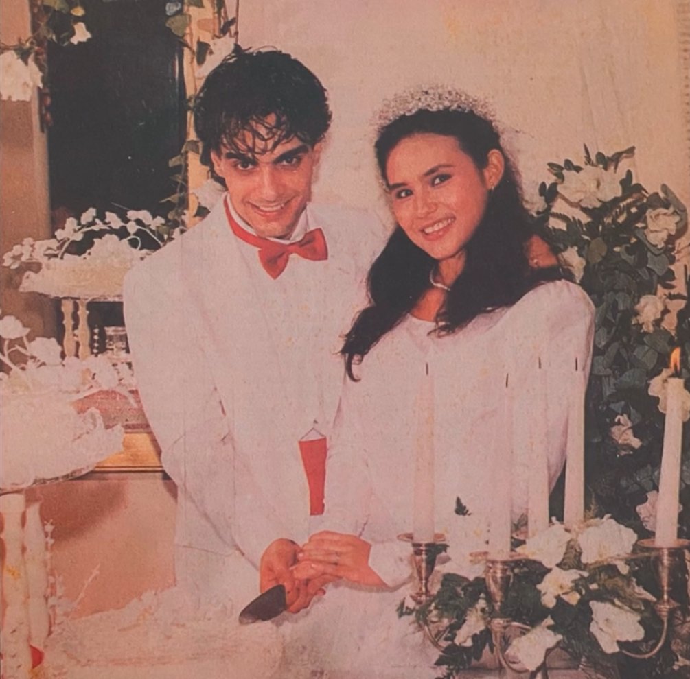 Na imagem vemos Guilherme de Pádua e Paula Thomaz sorrindo e ambos segurando a faca para cortar o bolo do seu casamento. Ele com um terno branco e gravata vermelha e ela com um vestido de noiva e uma coroa de flores branca na cabeça. 