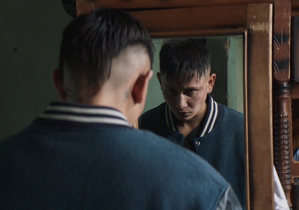 Cena do filme Um Homem. No centro da imagem, temos o ator Dylan Felipe Ramirez olhando sua reflexão em um espelho. Dylan é um jovem pardo, de cabelos pretos escuros, tatuagens perto das orelhas e olhos castanhos. Ele está vestindo uma jaqueta azul escura com detalhes em branco, em cima de uma camiseta também azul escura. O espelho é revestido de madeira marrom, provavelmente embutido a um guarda roupa da mesma cor. O cenário é uma parede esverdeada e desgastada. A cena acontece durante o dia. 