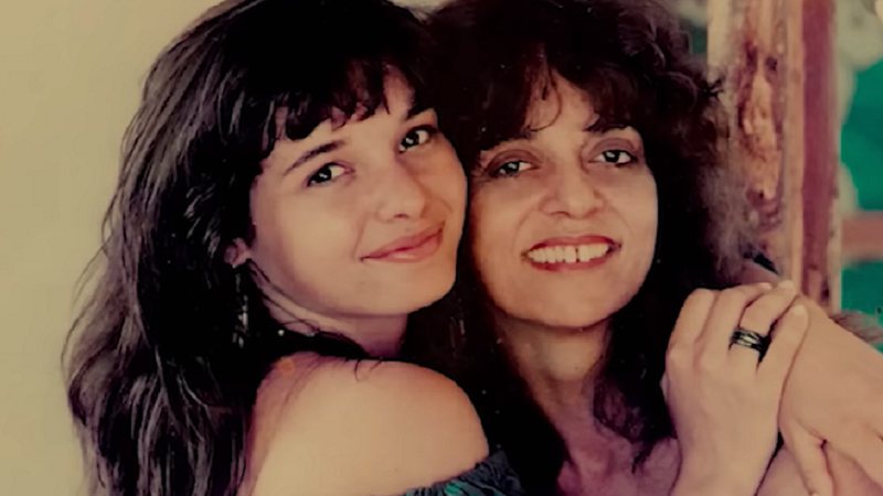 Na imagem vemos Daniella Perez ao lado de sua mãe, a novelista Glória Perez. As duas encontram-se abraçadas, sorrindo e olhando diretamente para a câmera. 