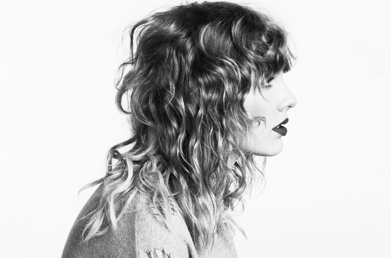 A imagem mostra Taylor Swift, mulher branca e loira. Ela está de perfil e tem os cabelos médios e cacheados, e usa um batom escuro. A foto é em preto e branco e não há nada ao fundo.