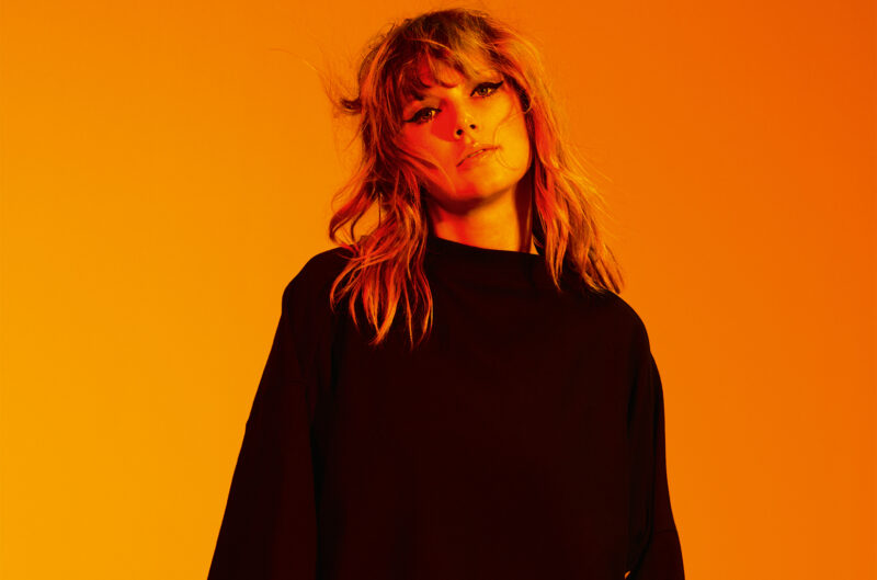 Taylor Swift, mulher branca e de cabelos claros, aparece ao centro da foto. Ela usa uma blusa larga, de mangas compridas e preta. O fundo é laranja. A foto inteira possui uma coloração alaranjada.