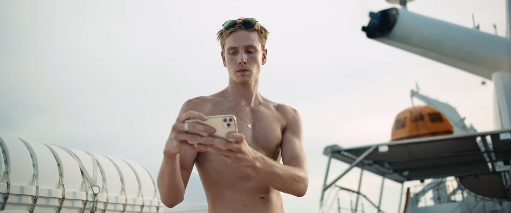 Cena do filme Triângulo da Tristeza, mostra um homem branco, sarado e sem camisa, tirando uma foto com um celular em posição horizontal. Ao fundo, vemos o céu e está de dia. 